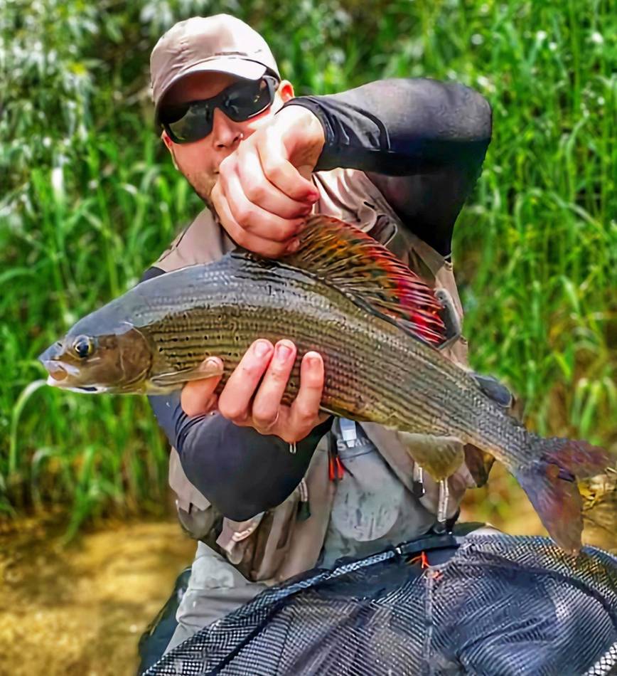 A guest smiling with his grayling by the riverbank