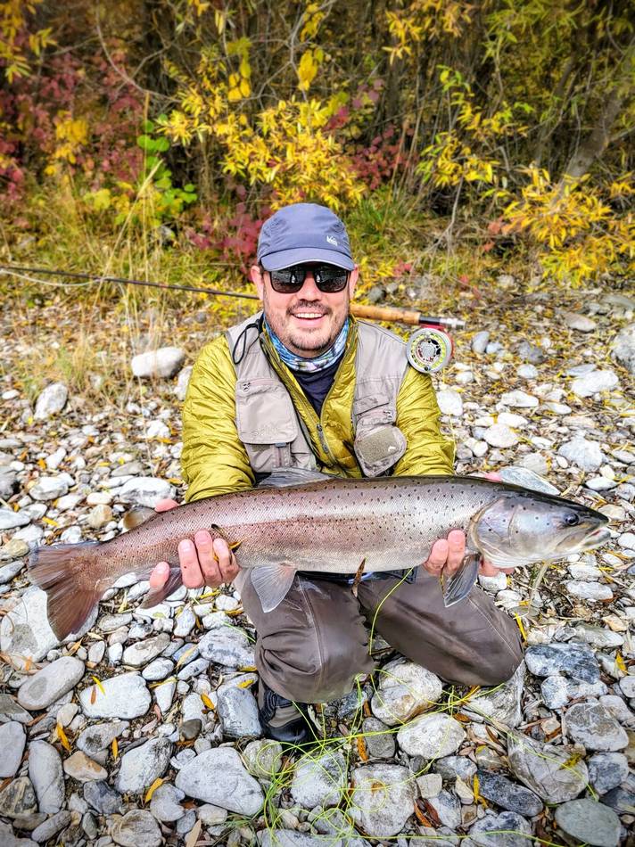 A guest holding a large silver trout on the riverbank in autumn